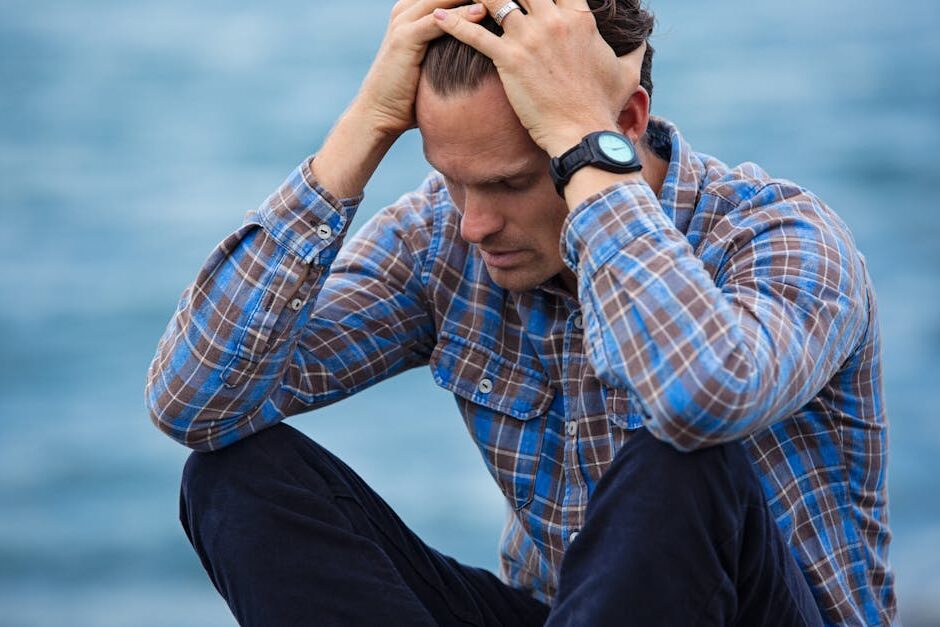 A man in a plaid shirt sits by the water looking distressed, symbolizing stress.